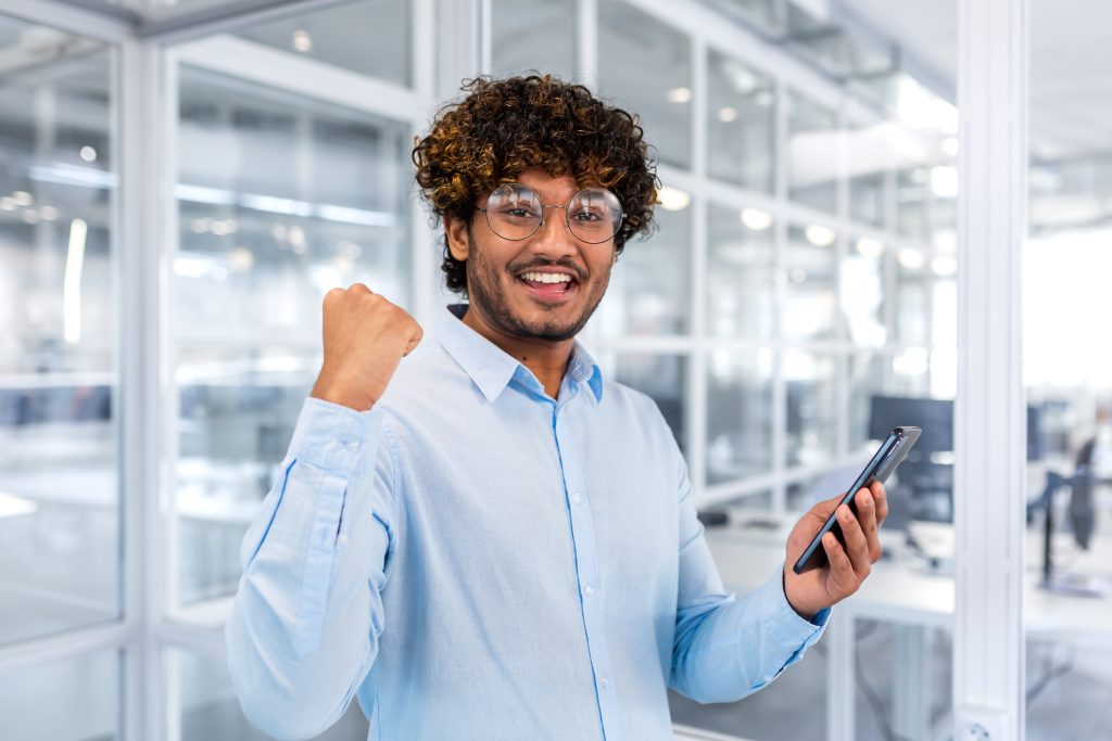 Profissional feliz em escritório moderno segurando celular e comemorando sucesso no ambiente de trabalho.